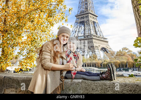 Herbst Kurzurlaube in Paris mit der Familie. Porträt von lächelnden Mutter und Tochter Reisende am Ufer in der Nähe von Eiffelturm in Paris, Frankreich, sitzen auf der Brüstung Stockfoto