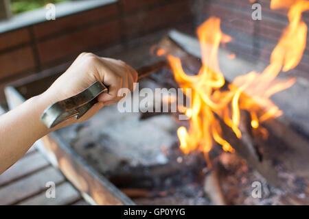 Starten Sie Feuer für Holzkohle-Grill. Arm mit langstieligen Zangen Einstellung Feuer Closeup. Helle Flammen und Brennholz. Stockfoto