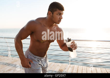Ernsthafte afroamerikanischen jungen Sportler laufen und halten Sie im freien Wasser Flasche Stockfoto