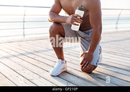 Zugeschnittenes Bild entspannt afrikanischen Sportler stretching und Flasche Wasser am Pier holding Stockfoto