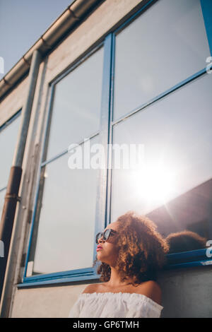 Schuss von schöne junge afrikanische Frau im Freien sitzen und wegsehen. Frau mit Sonnenbrille draußen zu sitzen. Stockfoto