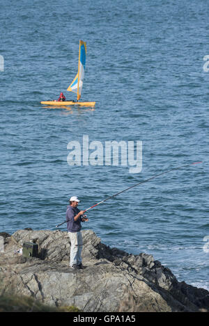 Ein Mann Fische aus Felsen als gelbe Trimaran Segel Vergangenheit im Hintergrund. Stockfoto