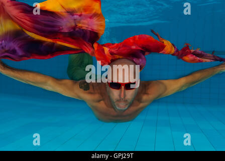 Männliches Modell fliegen unter Wasser trägt rote Sonnenbrille und hält ein buntes Tuch gegen das Blau des Swimmingpools Stockfoto
