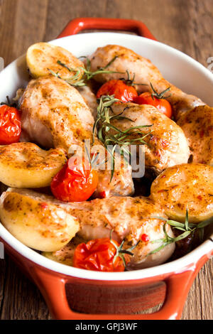 Ofen gebackene Hähnchenschenkel mit Gemüse (Tomaten, Kartoffeln), Kräuter und Gewürze in Auflaufform auf rustikalen hölzernen Hintergrund Stockfoto