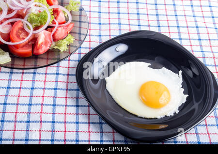 Nützliche kleine hausgemachtes Frühstück mit Spiegelei auf schwarzen Druckplatte und Gemüse auf einem karierten Tischdecke aus Stoff Stockfoto