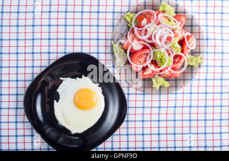 Draufsicht auf die Oberfläche des Tisches mit einer schwarzen Platte mit Spiegelei und einer kleinen Portion Tomatensalat auf einem Stoff-tablecl Stockfoto