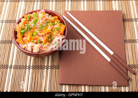 Draufsicht auf eine asiatische Schale Reisnudeln und Gemüse Gewürze in eine kleine Holzschale auf einer gestreiften Matte Stockfoto