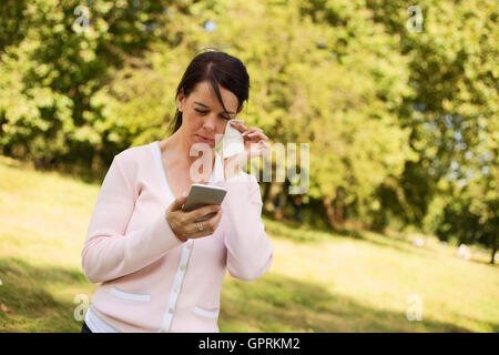 Frau eine Träne aus ihren Augen abwischen, da sie Soome schlechte Nachrichten auf ihr Handy liest. Stockfoto