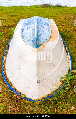 Upside-down Boot an Land. Das Boot hat blaue Kiel und Vergilben weiße Seiten. Trockene Blätter haben damit begonnen, um ihn herum zu sammeln. Stockfoto