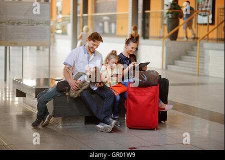 Junge Eltern mit zwei Kindern am Bahnhof. Familie in einem großen Wartesaal. Alle haben sich auf eine Bank niedergelassen. Dunkl Stockfoto