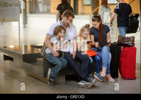 Junge Eltern mit zwei Kindern am Bahnhof. Familie in einem großen Wartesaal. Alle haben sich auf eine Bank niedergelassen. Dunkl Stockfoto