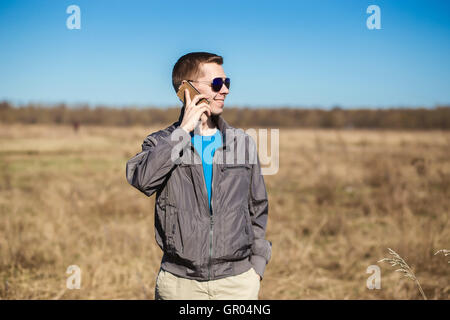 Junger Mann mit Sonnenbrille und Windbreaker telefonieren mit dem Handy. Es gibt ein Feld als Hintergrund Stockfoto