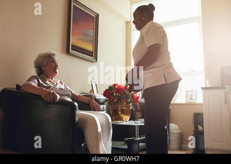 Ältere Frau sitzend auf einem Stuhl zu Hause mit weiblichen Betreuer stehen. Krankenschwester visiting senior Patienten zur Überprüfung blo Stockfoto