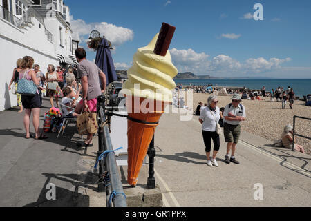 Ältere Paar zu Fuß entlang der Strandpromenade an einem Kunststoff-Eis-Kegel an der britischen Küste in Lyme Regis, Dorset England UK KATHY DEWITT Stockfoto