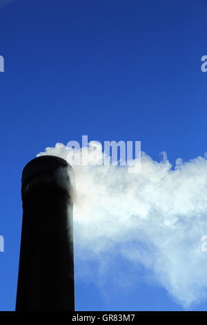 Rauch strömt aus einem Schornstein unter einem pulsierenden klaren blauen Himmel. CO2-Emissionen stehen ganz oben auf aktuelle Themen Bezug auf den Klimawandel Stockfoto