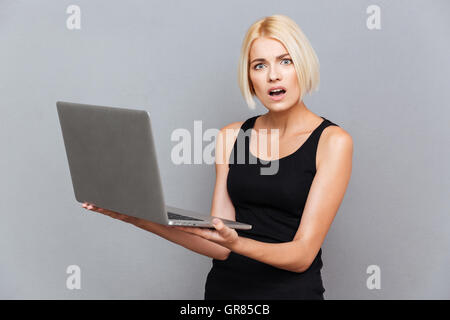 Unglückliche junge Frau mit Laptop über grauen Hintergrund erstaunt Stockfoto