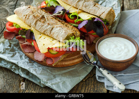 Leckere Sandwiches mit Schinken, Käse, Salat, Tomate und einer Schüssel saure Sahne auf einem hölzernen Hintergrund Stockfoto