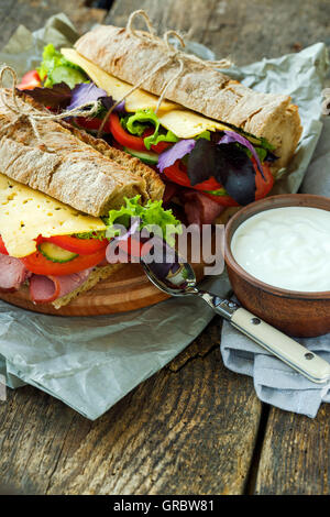 Leckere Sandwiches mit Schinken, Käse, Salat, Tomate und einer Schüssel saure Sahne auf einem hölzernen Hintergrund Stockfoto