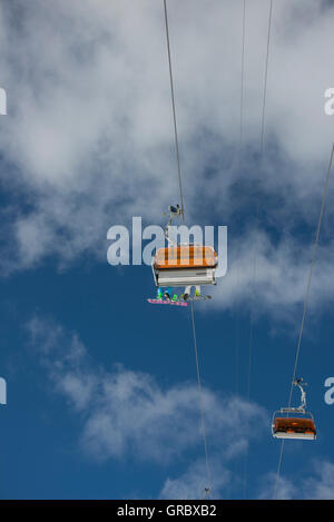 Sesselbahn mit Orangen Wetterschutzhauben und zwei Snowoarders zeigt die Unterseite ihrer Platten, gegen den blauen Himmel und weiße Wolken Stockfoto