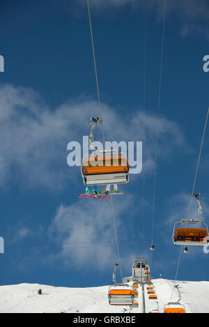 Sesselbahn mit Orangen Wetterschutzhauben und zwei Snowoarders zeigt die Unterseite ihrer Platten, gegen den blauen Himmel und weiße Wolken Stockfoto