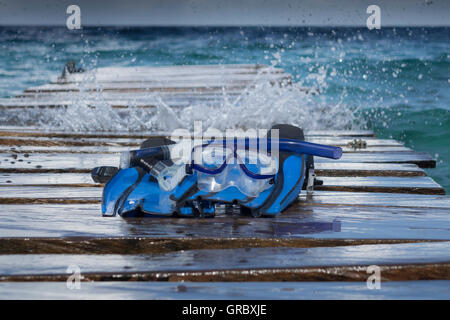 Maske, Schnorchel und Flossen am Holzsteg, im Hintergrund Sprays von Wasser und Meer In Bewegung Stockfoto