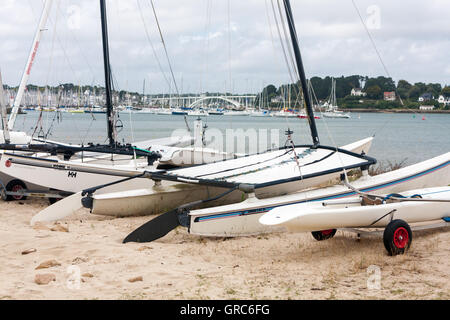 Eine Reihe von Booten und beachable Katamarane auf dem Sand in La Trinité-Sur-Mer, Bretagne, Frankreich, mit der Marina im Hintergrund Stockfoto