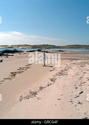 Frau zu Fuß auf der sandigen Strand von The Strand auf Insel Colonsay, mit der Insel Oronsay auf Distanz, Schottland, UK. Stockfoto