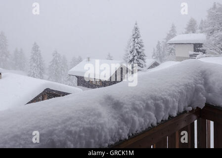 Schnee fällt. In den Vordergrund verschneite Holzgeländer, im Hintergrund Bäume einige Chalets und Tanne In einer winterlichen Landschaft Stockfoto