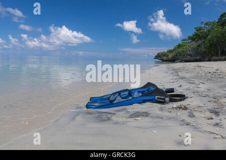 Blaue Flossen, Maske und Schnorchel auf einem weißen Sandstrand. Im Hintergrund Flachwasser, blauer Himmel, weiße Wolken und grüne Vegetation Stockfoto