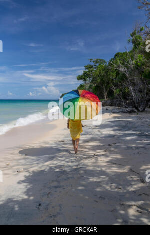 Frau In einem gelben Kleid und einen Regenbogen farbige Sonnenschirm Spaziergänge an einem Sandstrand. Stockfoto