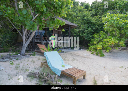 Weibliche Schnorchler vor Holzbungalow, umgeben von üppigen Buschwerk, In den Vordergrund Liegestuhl am Sandstrand Stockfoto