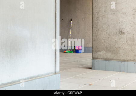 Reinigungsgeräte (Besen, Eimer, Lappen und Handschuhe) vor einem grauen, monotonen Betonwand und verschmutzten Boden bunt. Stockfoto