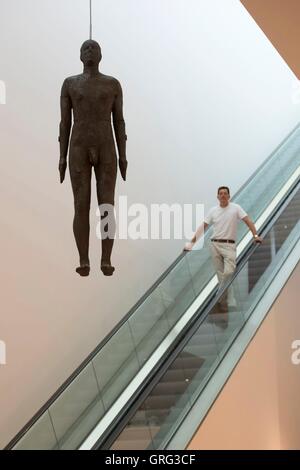 Antony Gormley reist auf einer Rolltreppe vorbei eine lebensgroße Skulptur der selbst - Objekt, 1999 - die hängt von der Decke auf den Ondaatje Wing Main Hall von der National Portrait Gallery, London. Stockfoto