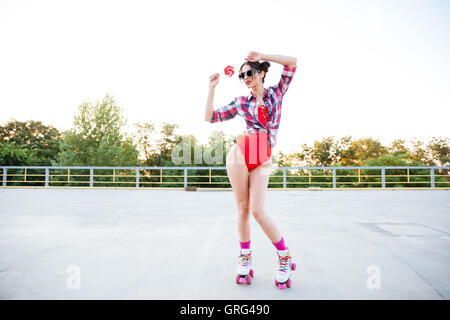 Schöne junge Frau im roten Badeanzug und kariertes Hemd holding Lutscher und stehen auf Rollschuhen Stockfoto