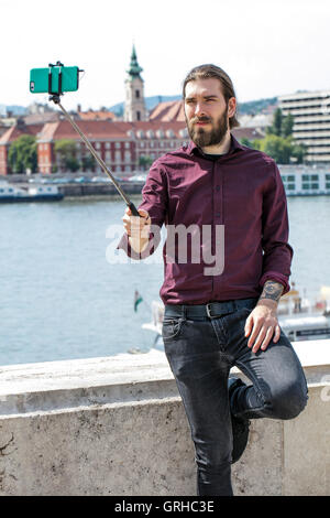 Eine junge Hipster steht unter einem Selfie auf einem Selfie Stock mit Blick auf die Donau rive r in Budapest Stockfoto