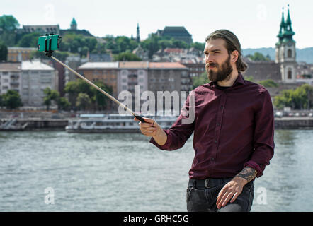 Eine junge Hipster steht unter einem Selfie auf einem Selfie Stock mit Blick auf die Donau rive r in Budapest Stockfoto