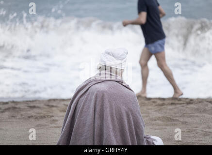 Älterer Mann mit Turban am Strand mit Blick auf den Ozean mit Jogger im Hintergrund Stockfoto