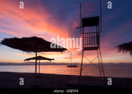 Benitses Korfu Griechenland Sonnenaufgang am Strand mit Stroh Sonnenschirme, Boot und Rettungsschwimmer Turm Silhouette Stockfoto