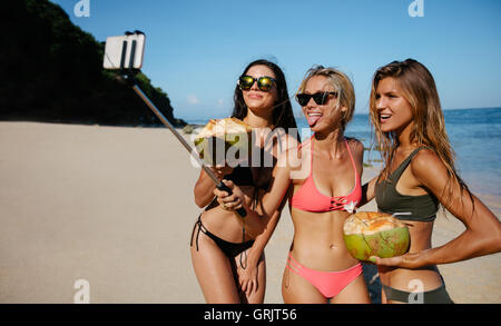 Drei junge Frauen im Bikini am Strand posieren für Selbstporträt. Gruppe von Freundinnen halten Kokosnüsse und nehmen Selfie wi Stockfoto