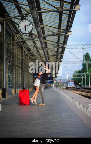 Paar steht auf der leeren Plattform unter der Uhr. Junger Mann umarmt und küsst eine Mädchen. Neben ihnen, einen großen roten Koffer. Ich Stockfoto