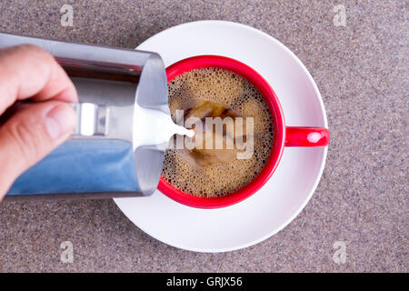 Man gießt Milch aus einem Krug in eine Tasse frisch gebrühten schwarzen Filter oder Espresso Kaffee in einem roten Becher auf eine weiße Untertasse gegen Stockfoto