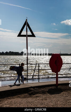 Ein kleines Mädchen mit einem Surfbrett, Silhouette gegen die rauen Wasser des Flusses Colne, Brightlingsea, Essex, UK Stockfoto