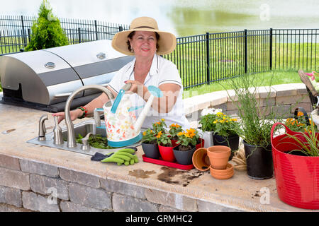 Freundliche attraktive ältere Dame in einen breitkrempigen Stroh Sonnenhut Bewässerung ihrer Zimmerpflanzen auf eine Terrasse, die Füllung der Dose Stockfoto