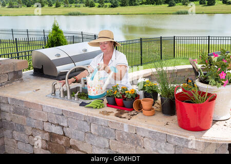 Senior Lady Ausfüllen einer Gießkanne an eine Spüle in eine Sommerküche auf einer Terrasse zu ihrer Sammlung von Topfpflanzen Wasser ein Stockfoto