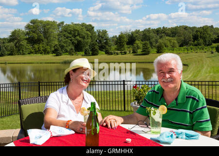 Liebevolle senior Brautpaar sitzt an einem Gartentisch auf eine Terrasse mit Blick auf einen ruhigen See in einer üppigen Landschaft s Stockfoto