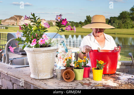 Ältere Dame in einen breitkrempigen Sonnenhut Stroh tendenziell ihre Topfpflanzen auf eine Terrasse mit Blick auf einen See als She transplan Stockfoto