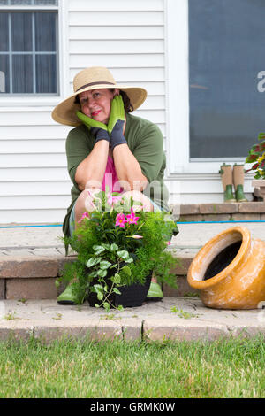 Stilvolle Oma in Gummistiefel und Sonnenhut sitzt auf dem Brick Schritte von ihrer Terrasse schaut nachdenklich in die Kamera mit einem schönen smil Stockfoto