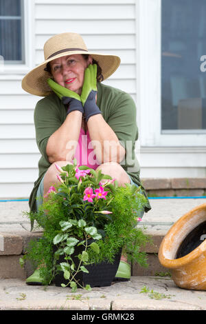 Attraktive senior Frau in einem stilvollen breitkrempigen Sonnenhut Stroh und Gartenhandschuhe Blumenerde Pflanzen auf ihrer Terrasse Schritten anhalten Stockfoto