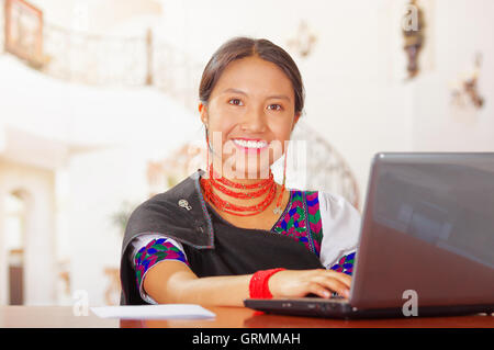 Junge Brünette tragen einheimische Tracht als Rezeptionist mit freundlichen Lächeln, Gesichtspunkt mit Computer, Kunden Stockfoto