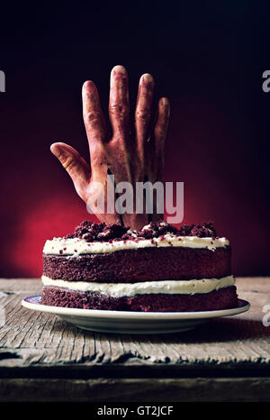 ein roter Samt-Kuchen gekrönt mit einer blutigen Hand in eine gruselige Szene für Halloween, auf einem rustikalen Holztisch Stockfoto